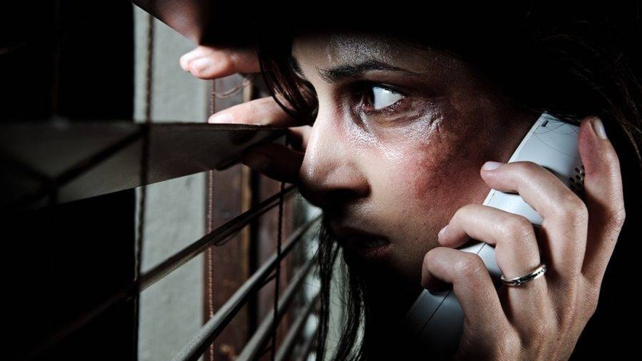 Fearful battered woman peeking through the blinds to see if her husband is home while calling for help